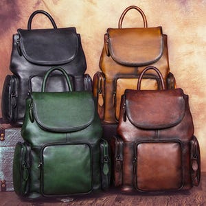 May include: Four leather backpacks in black, tan, dark green, and brown. Each bag features a top handle, a fold-over flap with a secure closure, a large front pocket, and two side pockets with zippers. The leather has a smooth, polished finish.