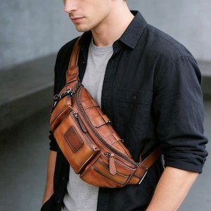 Puede incluir: Un bolso bandolera de cuero color canela con un compartimento principal con cremallera, un bolsillo frontal más pequeño con cremallera y un detalle de parche rectangular. El bolso tiene un diseño estructurado con costuras visibles y herrajes de metal oscuro.