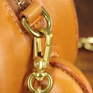 May include: Close-up of a tan leather bag with a gold-colored metal clasp and zipper. The bag's leather has a smooth texture, and the metal hardware is shiny. A gold chain is visible in the background.