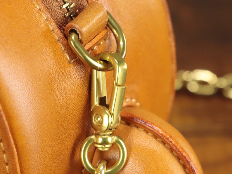 May include: Close-up of a tan leather bag with a gold-colored metal clasp and zipper. The bag's leather has a smooth texture, and the metal hardware is shiny. A gold chain is visible in the background.