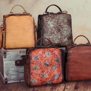 May include: A collection of leather handbags in different styles and colours. The bags include a tan backpack, a dark green and brown satchel, a red and blue floral patterned bag, and a brown satchel. They are displayed on a wooden surface.