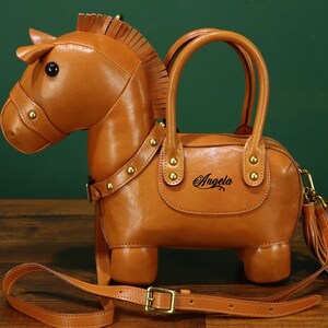 May include: A brown leather handbag shaped like a horse, featuring a head, body, and four legs. The bag has a top handle, a shoulder strap, and a decorative tassel. The name "Angela" is written on the side.