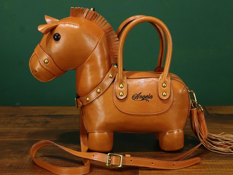 May include: A brown leather handbag shaped like a horse, featuring a head, body, and four legs. The bag has a top handle, a shoulder strap, and a decorative tassel. The name "Angela" is written on the side.