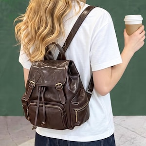 Puede incluir: Una mochila de cuero marrón oscuro que presenta una solapa superior con dos cierres de hebilla, un compartimento principal con cordón y tres bolsillos exteriores con cremallera. Incluye correas ajustables y herrajes de metal dorado.
