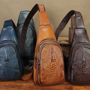 May include: Four leather sling bags in shades of blue, green, tan, and brown. Each bag features a textured, crocodile-like pattern and a single shoulder strap. The bags are displayed on a wooden surface.