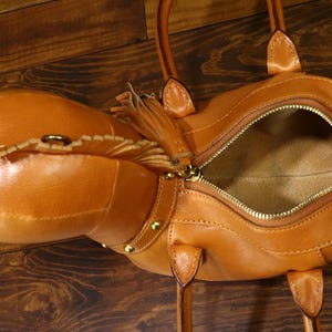 May include: A tan leather handbag with a horse head design. The bag has a zippered opening, gold-colored hardware, and a decorative tassel. The bag has two handles and a shoulder strap. The bag is made of leather.