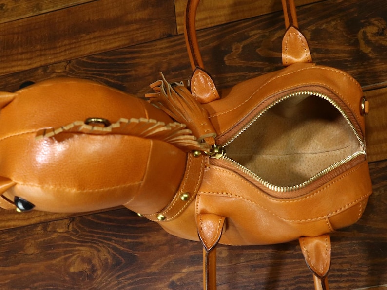 May include: A tan leather handbag with a horse head design. The bag has a zippered opening, gold-colored hardware, and a decorative tassel. The bag has two handles and a shoulder strap. The bag is made of leather.