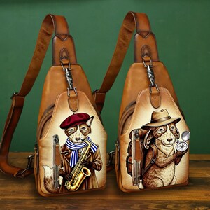 Puede incluir: Dos bandoleras de cuero marrón con ilustraciones de perros de dibujos animados. Uno lleva boina y toca el saxofón, el otro lleva sombrero y sostiene una brújula. Cada bolso tiene una correa de cuero marrón y herrajes plateados.
