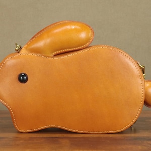 May include: A tan leather bunny-shaped handbag with a black eye and a small, round tail. The bag has a zipper closure and a small handle. The bag is made of smooth leather and has a stitched design.
