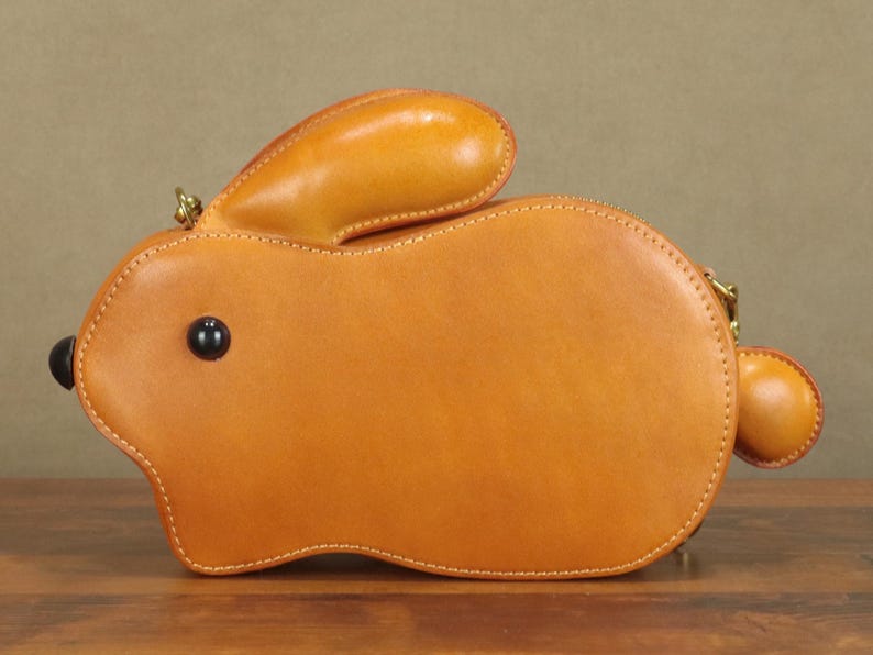 May include: A tan leather bunny-shaped handbag with a black eye and a small, round tail. The bag has a zipper closure and a small handle. The bag is made of smooth leather and has a stitched design.