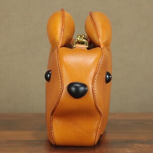 May include: A tan leather handbag shaped like a bear's head, featuring black eyes and a nose. The bag has two rounded ears at the top and a gold-colored chain. The stitching is visible, and the bag appears to be sitting on a wooden surface.
