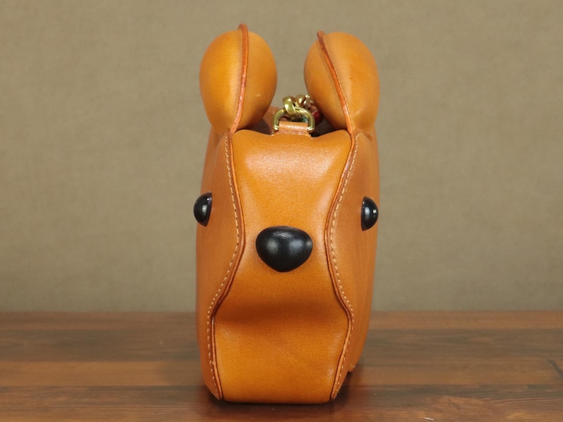 May include: A tan leather handbag shaped like a bear's head, featuring black eyes and a nose. The bag has two rounded ears at the top and a gold-colored chain. The stitching is visible, and the bag appears to be sitting on a wooden surface.