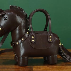 May include: A brown leather handbag shaped like a horse. The bag has a handle, a shoulder strap, and a tassel. Gold-colored studs adorn the collar and the bag's body. The horse's head has a bridle and a small horn.