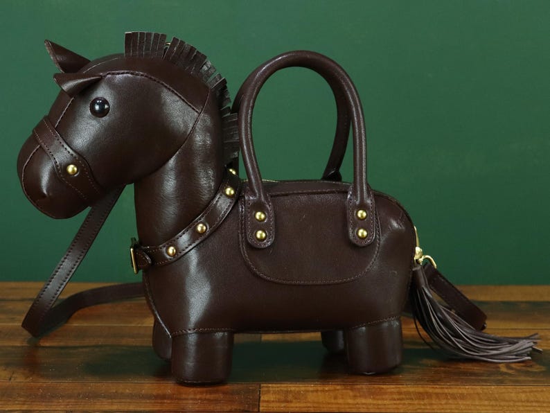 May include: A brown leather handbag shaped like a horse. The bag has a handle, a shoulder strap, and a tassel. Gold-colored studs adorn the collar and the bag's body. The horse's head has a bridle and a small horn.