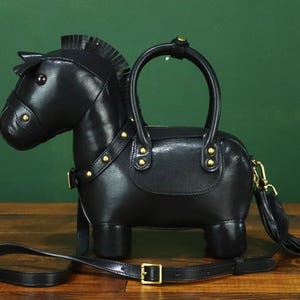 May include: Black leather horse-shaped handbag with a top handle and a detachable shoulder strap. The bag features gold-colored hardware, including studs and a buckle. A tassel hangs from the side.