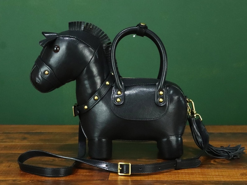 May include: Black leather horse-shaped handbag with a top handle and a detachable shoulder strap. The bag features gold-colored hardware, including studs and a buckle. A tassel hangs from the side.