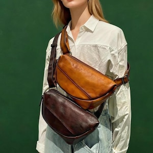 Puede incluir: Dos bolsos cruzados en forma de media luna que se llevan sobre el hombro. El bolso superior es de cuero color canela cálido, mientras que el inferior es de cuero marrón oscuro. Ambos bolsos tienen un acabado liso, silueta curva y correas ajustables.