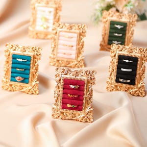 May include: Several ornate gold picture frames displaying rings. Each frame has a velvet insert in various colours, including teal, pink, green, red, and black. The rings are of different styles and colours, set against the velvet.