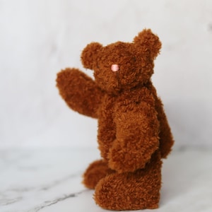 May include: A small, fuzzy brown teddy bear with a textured, plush surface. The bear features rounded ears, a small pink nose, and is posed with one arm raised. The material appears to be a soft, curly synthetic fabric, giving the toy a distinct, tactile appearance throughout its entire body.