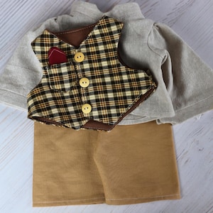 Könnte beinhalten: Ein Puppenoutfit bestehend aus einem hellgrauen Langarmshirt, einer braun-beigen Weste mit Karomuster, drei gelben Knöpfen und einem kleinen dunkelroten Einstecktuch sowie einer beigen Shorts. Die Kleidungsstücke liegen übereinander.