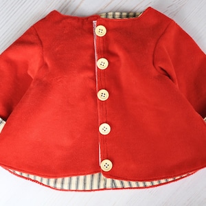 May include: A bright red long-sleeved jacket featuring a front button closure with five light-colored circular buttons. The garment has a flared hem and sleeve cuffs that reveal a black and white striped pattern on the interior lining.