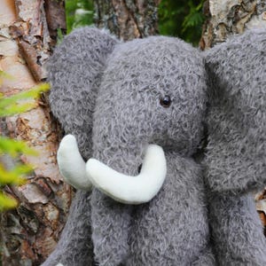 Op de afbeelding: Een pluche olifant gemaakt van gestructureerd grijs imitatiebont. Het knuffeldier heeft grote, slappe oren, een prominente slurf en twee gladde, gebogen witte slagtanden die uit de basis van de slurf steken. Het heeft donkere ogen.