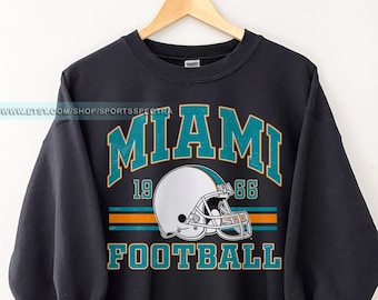 May include: Black sweatshirt with a vintage-style Miami Dolphins logo. The logo features a teal and orange football helmet with the text "Miami" in teal and "1966" in orange above the helmet. The text "Football" is in yellow below the helmet.
