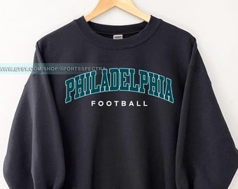 May include: Black sweatshirt with the word "PHILADELPHIA" in teal arched across the chest, with "FOOTBALL" in white below. The sweatshirt is hanging on a wooden hanger.