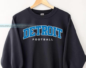 May include: Black sweatshirt with the word "DETROIT" in blue block letters with a white outline, above the word "FOOTBALL" in white, displayed on a wooden hanger. The sweatshirt is a crew neck style.