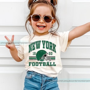 May include: A toddler wearing a white t-shirt with a green and white graphic that says "New York 19 60 Football" with a football helmet graphic. The toddler is wearing blue denim shorts and sunglasses.