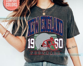 May include: A gray t-shirt with a vintage-style graphic design featuring the words "New England" in blue and red, "1960" in white, and "Football" in white. The graphic also includes a gray and red football helmet.
