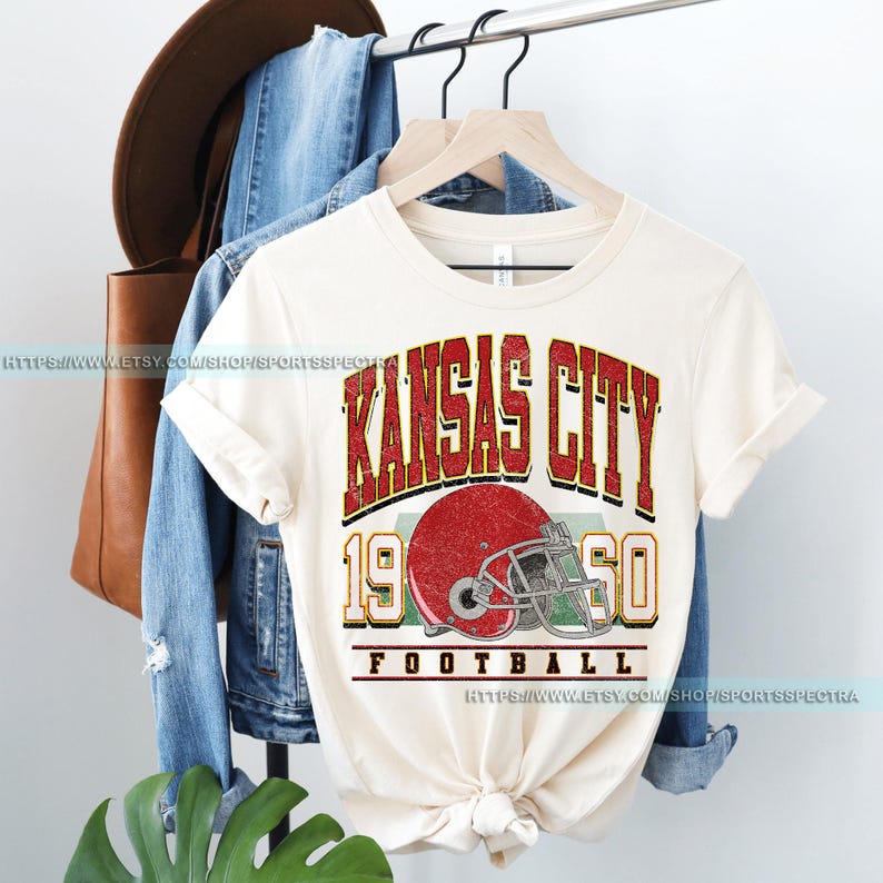 May include: A cream-colored t-shirt with a vintage-style graphic design featuring the text "Kansas City 1960 Football" and a red and white football helmet.