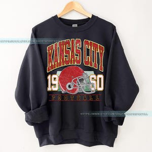 May include: Black sweatshirt with a vintage-style graphic of the Kansas City Chiefs logo, featuring a red helmet and the text "Kansas City 1960 Football".