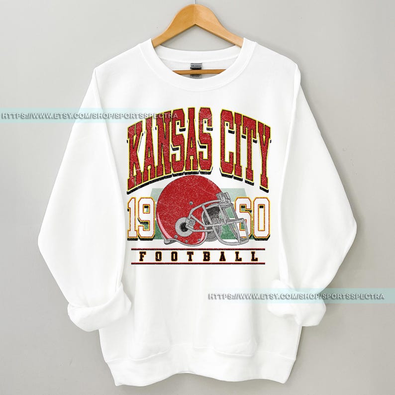May include: White sweatshirt with a vintage-style graphic design featuring the text "Kansas City 1960 Football" and a red football helmet.