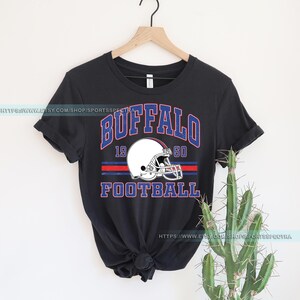 Może przedstawiać: Czarny T-shirt z grafiką w stylu vintage przedstawiającą kask do futbolu i tekst "Buffalo 19 60 Football".