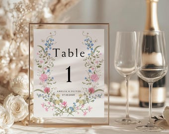 May include: A gold-framed table number card with a floral design in shades of pink, blue, and green. The card reads "Table 1 Amelia & Oliver 07.09.2026".