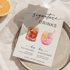 May include: A drinks menu card with the words "Signature Drinks" and two cocktail illustrations. The "Old Fashioned" cocktail is in a rocks glass, and the "Blushing Bride" is in a stemless glass. The card includes drink ingredients and a wedding date.