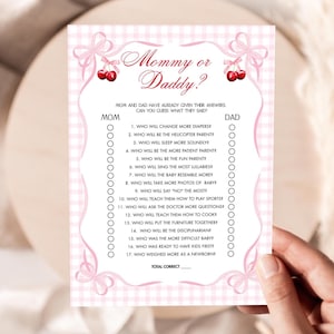 May include: A pink and white baby shower game card with the text "Mommy or Daddy?". The card features a gingham pattern, decorative bows, and a list of questions to guess who will do what. The card is held by a person.