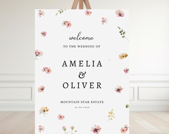Op de afbeelding: Een wit welkomstbord voor een bruiloft op een houten ezel. Het bord bevat de woorden "welcome" en "to the wedding of Amelia & Oliver" in zwarte tekst. Delicaat geschilderde bloemen in roze, geel en groen zijn verspreid.
