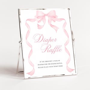 K&ouml;nnte beinhalten: Ein gerahmtes Schild mit den Worten "Diaper Raffle" in rosa Schrift, verziert mit einer Aquarellschleife und B&auml;ndern. Auf dem Schild steht: "Wenn Sie eine Packung Windeln f&uuml;r die Windel-Tombola mitgebracht haben, legen Sie bitte Ihr Ticket hier ab."