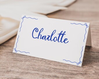 Pode incluir: Um cartão de lugar branco com o nome "Charlotte" em escrita azul. O cartão tem uma borda decorativa azul e está sobre uma superfície de madeira. O cartão é dobrado para ficar em pé.