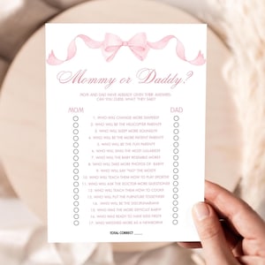 May include: A white paper game card with the text "Mommy or Daddy?" in pink script, featuring a pink bow and ribbon design. The card lists questions for a baby shower game, with spaces for Mom and Dad answers.