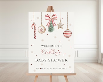 May include: A cream-colored sign on a wooden easel with Christmas ornaments and the text "Welcome to Emilly's Baby Shower." The ornaments include a candy cane, baubles, a bow, a star, and a small Christmas tree.