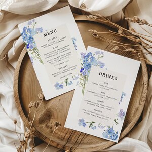 May include: Two white menu cards with blue floral accents, featuring the words "MENU" and "DRINKS." The cards are on a wooden tray with dried floral elements. The menu includes starters, main courses, desserts, and drinks.