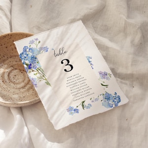 May include: A white table card with blue floral watercolor accents and the word "table" above the number "3". The names of guests are listed below the number. The card is placed on a white linen surface.