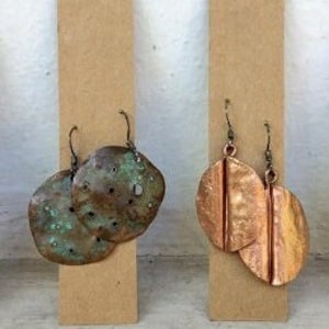 Könnte beinhalten: Zwei Paar handgefertigte Kupferohrringe, präsentiert auf Kartonständern. Ein Paar hat eine strukturierte, grünspanfarbene Oberfläche, das andere ein glattes, poliertes Kupferaussehen. Beide Paare haben einfache Hakenverschlüsse.