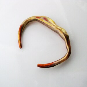 May include: A gold-toned metal bracelet with an organic, wavy shape. The bracelet has a reddish-brown hue on the inside.