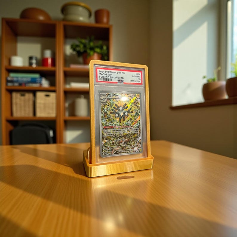 May include: A graded Pok&eacute;mon card in a yellow holder, displayed on a wooden table. The card features a colorful illustration and text. The holder is supported by a matching yellow stand. The background includes a wooden shelf with various items.