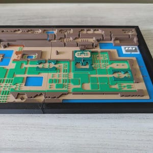 May include: A 3D-printed map of a video game world, featuring a detailed layout with green, brown, and blue sections. The map is set within a black frame, showcasing the intricate design and terrain features. The map is a unique piece of gaming decor.