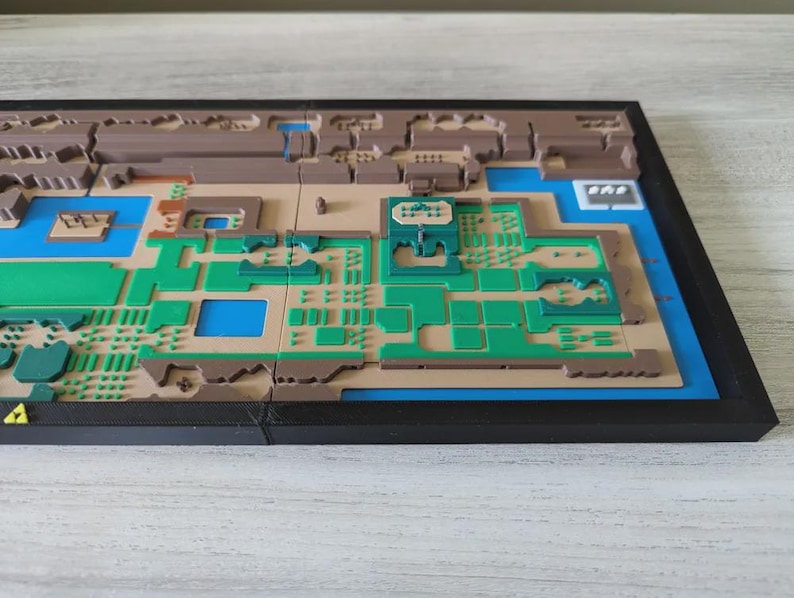 May include: A 3D-printed map of a video game world, featuring a detailed layout with green, brown, and blue sections. The map is set within a black frame, showcasing the intricate design and terrain features. The map is a unique piece of gaming decor.